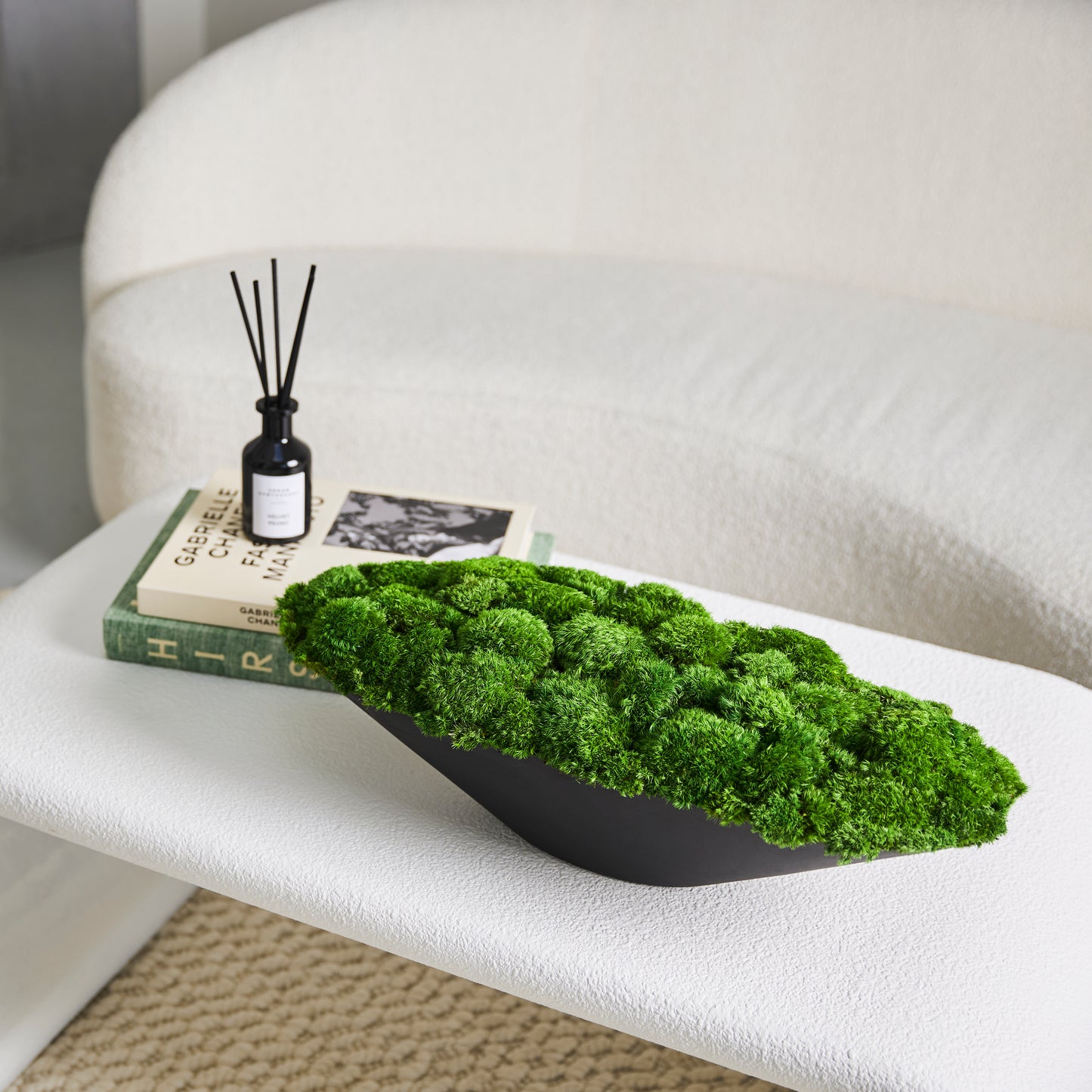 A lush piece of preserved moss. Finished with a black ceramic boat-shaped base. Displayed on a white coffee table.