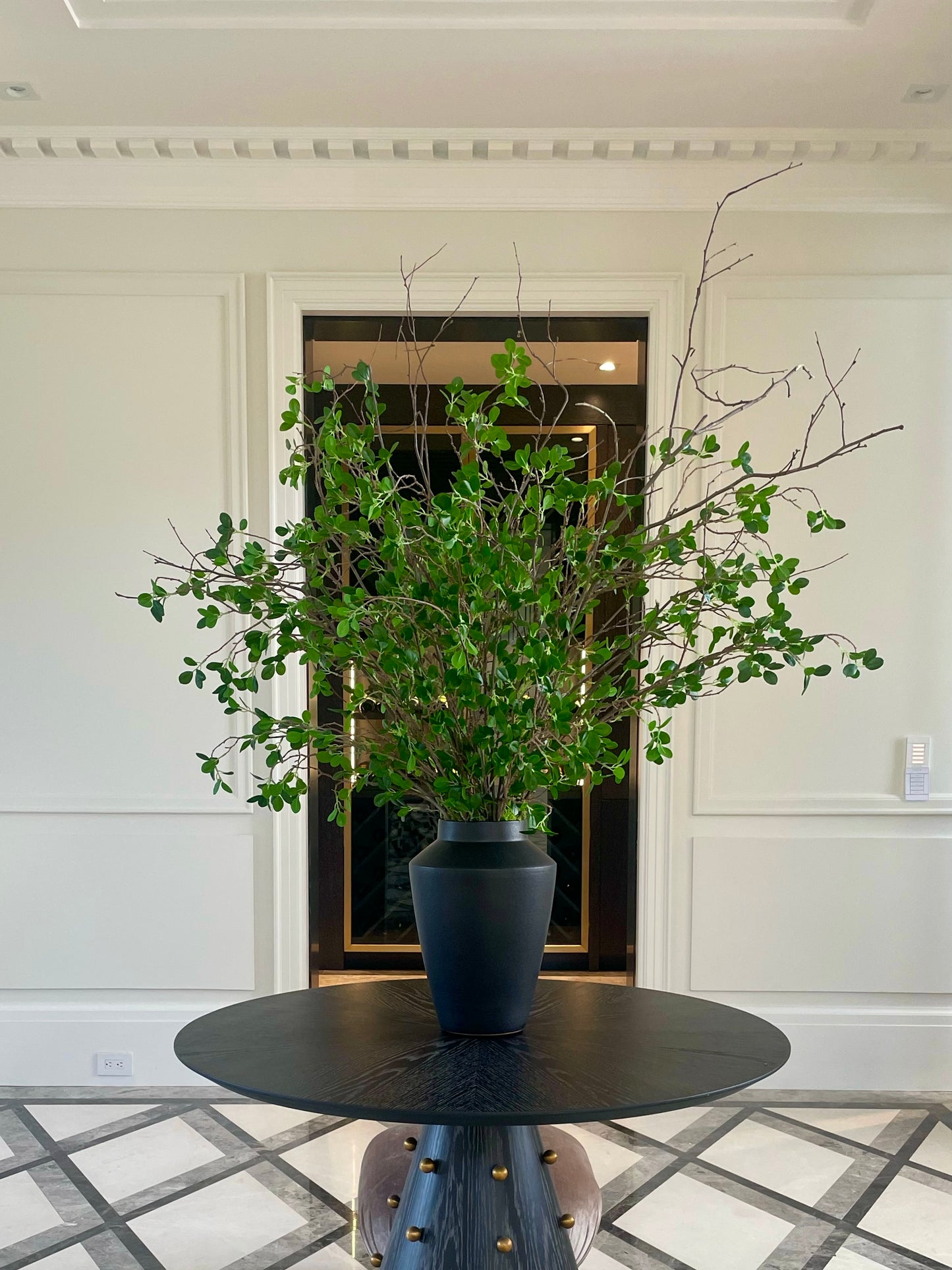 Natural looking ficus piece includes twig branch accents in a black ceramic vase. Displayed in a front hall way of a home.